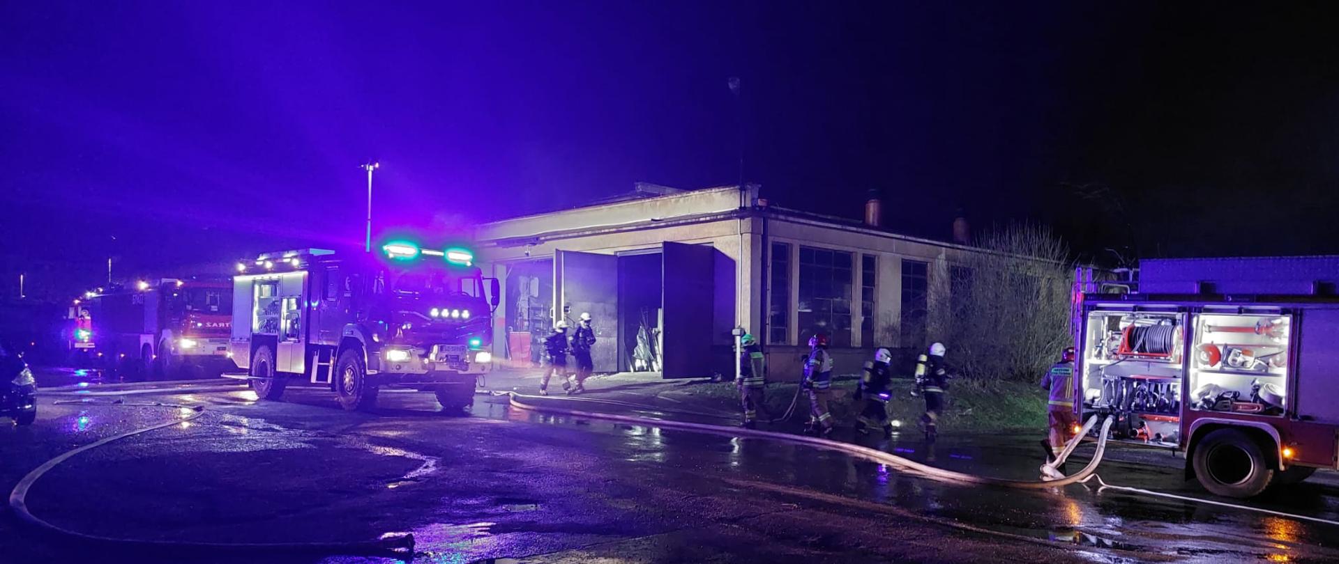 Ustawione samochody strażackie zabezpieczają miejsce zdarzenia i przy użyciu masztów oświetleniowych na dachach oświetlają teren pożaru. Z samochodów rozwinięte na jezdni są węże pożarnicze. Strażacy prowadzą działania gaśnicze.