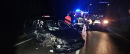 Samochód osobowy z uszkodzonym przodem na jezdni w tle służby ratownicze. Pora nocna.