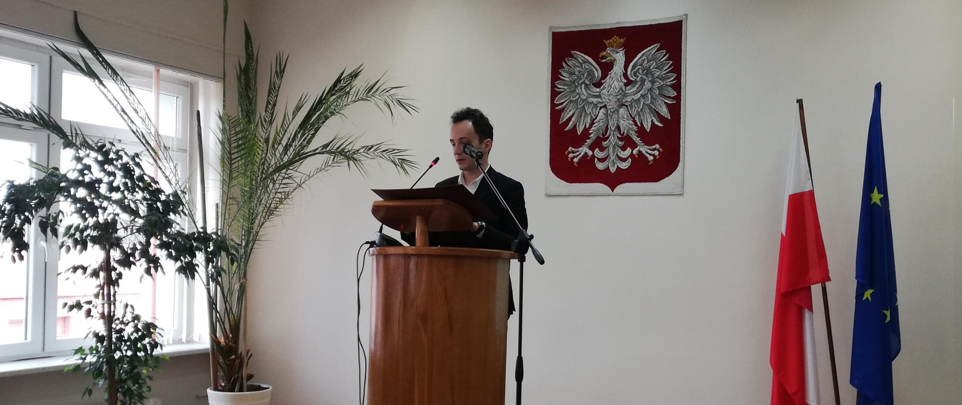 Na zdjęciu mężczyzna stoi przy mównicy. W tle flaga Polski oraz Unii Europejskiej 