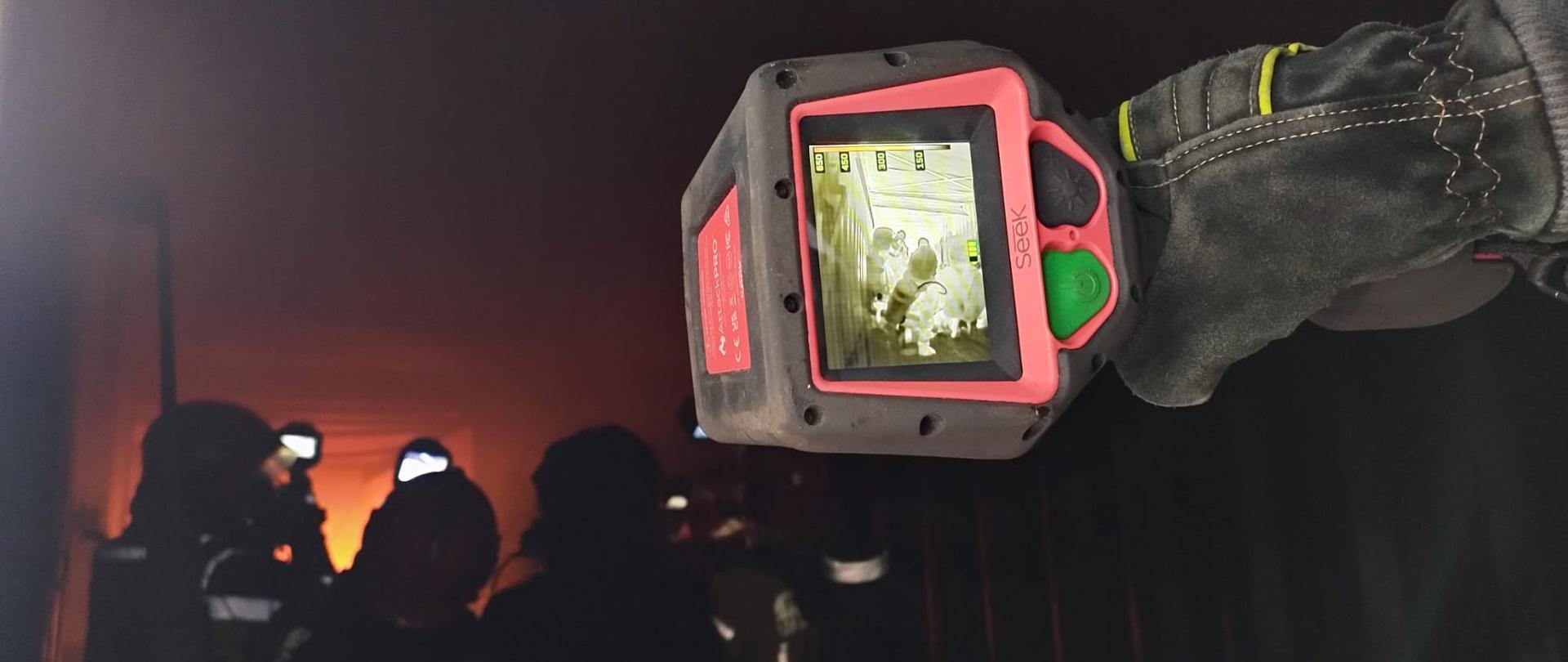 Ręka w rękawicy strażackiej trzyma kamerę termowizyjną, skierowaną na sylwetki innych strażaków i pożar w tle.