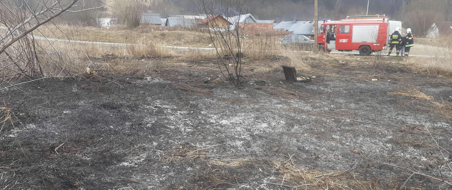 Spalona połać trawy i opona. W tle stoi samochód pożarniczy oraz strażacy.
