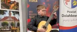 Na taborecie siedzi chłopiec ubrany w czarną koszulę oraz czarne spodnie i gra na gitarze. Za nim duże plakaty reklamujące powiat działdowski oraz konkurs gitarowy.