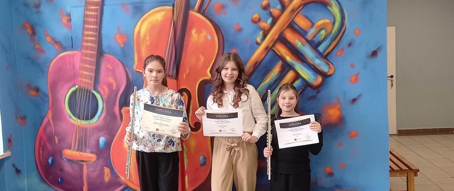 Trzy uczennice z dyplomami na tle muralu w szkole muzycznej przedstawiającego instrumenty muzyczne, gitara, skrzypce i trąbka.