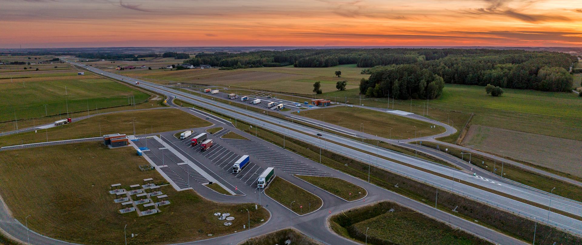 Droga ekspresowa widziana z góry, po jej obu stronach Miejsca Obsługi Podróżnych, czyli miejsca parkingowe dla samochodów, budynek z toaletami, miejsca do odpoczynku. 