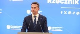 Rzecznik rządu, Minister Adam Szłapka podczas konferencji prasowej 01.07.2025 r. W tle ekran rzecznika rządu z logo Kancelarii Prezesa Rady Ministrów i mediami społecznościowymi rzecznika.