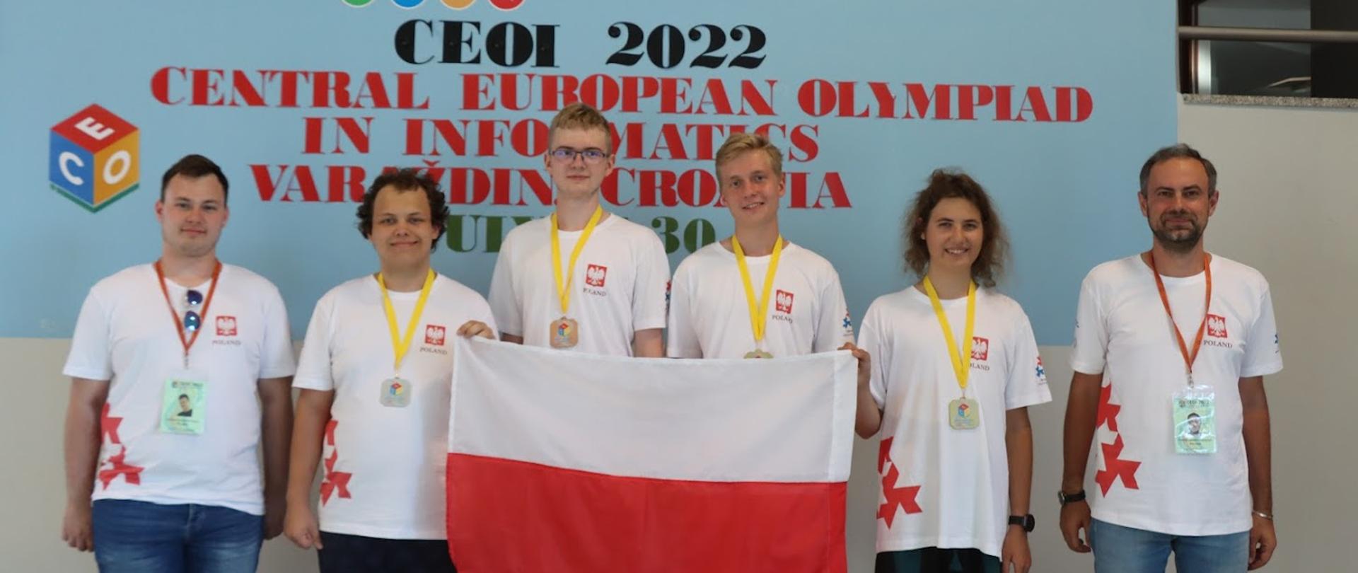 Sześcioro młodych ludzi stoi pod ścianą z napisem CEOI 2022, dwaj trzymają polską flagę.