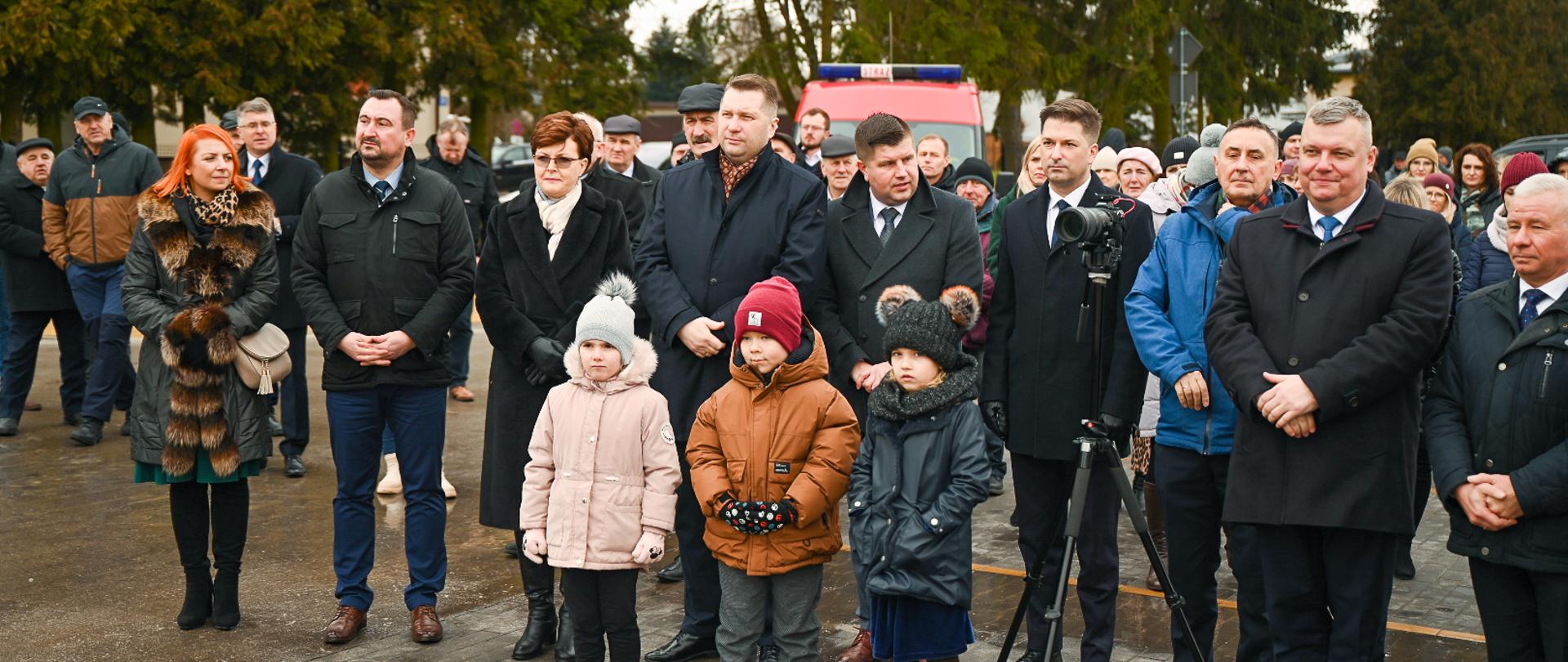 Zdjęcie na dworze, minister Czarnek stoi w dużej grupie ludzi, z przodu stoi troje małych dzieci, obok nich kamera na statywie, z tyłu drzewa.
