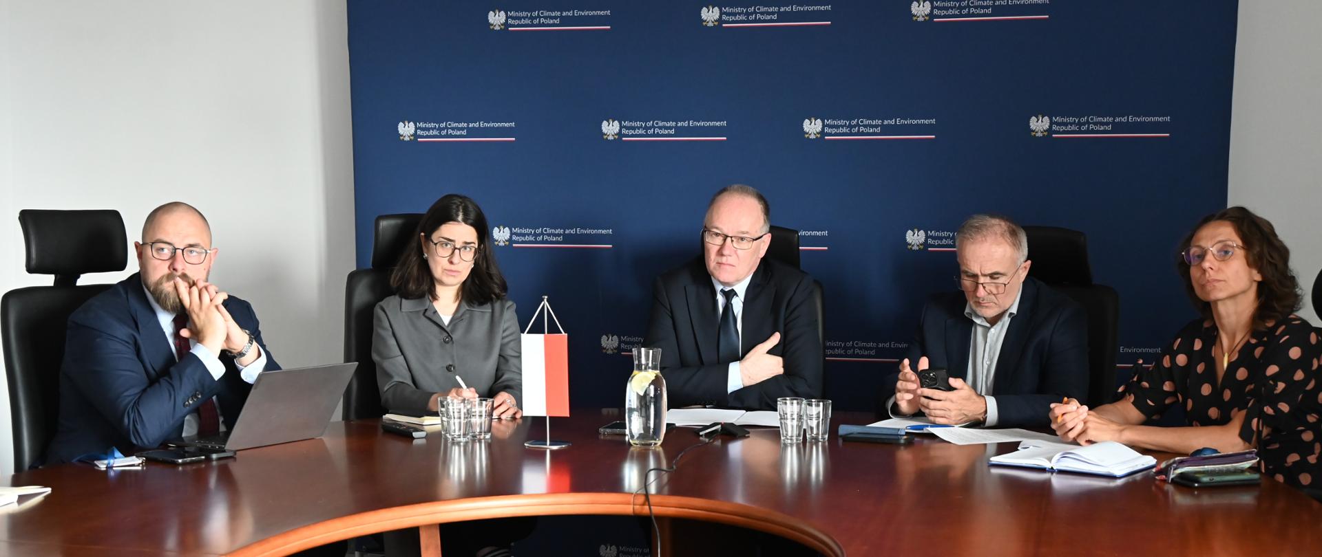 Wiceminister Krzysztof Galos, Główny Geolog Kraju, przewodniczył spotkaniu przedstawicieli służb geologicznych Polski i Kanady. Wiceminister siedzi w środku za okrągłym stołem, obok niego od prawej strony siedzą dwie osoby i od lewej jego strony dwie osoby. Wszyscy patrzą przed siebie, słuchają. Za osobami przy stole widać ściankę z granatowym tłem i logotypami Ministerstwa Klimatu i Środowiska.
