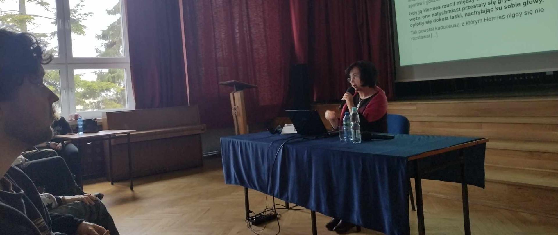 Na dużej auli, przed sceną, zwrócona do niej plecami, przy stole siedzi kobieta, trzyma mikrofon. W tle duży ekran z wyświetlonym podglądem ekranu. Przed kobietą widać rząd siedzących osób, zwróconych w jej stronę