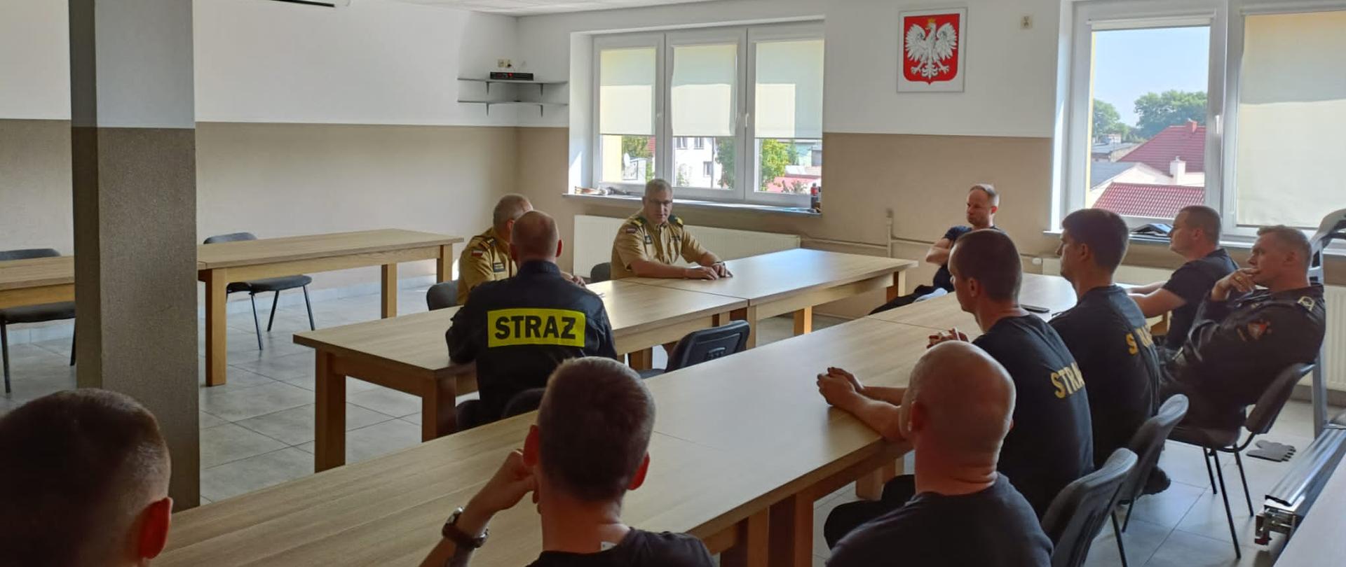 Strażacy siedzą przy stołach, rozmawiają z Kujawsko-Pomorskim Komendantem Wojewódzkim Państwowej Straży Pożarnej. Po prawej stronie ściana z 2 oknami a między nimi na ścianie wisi godło Polski.