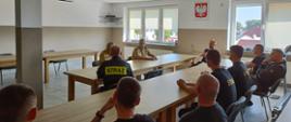 Strażacy siedzą przy stołach, rozmawiają z Kujawsko-Pomorskim Komendantem Wojewódzkim Państwowej Straży Pożarnej. Po prawej stronie ściana z 2 oknami a między nimi na ścianie wisi godło Polski.