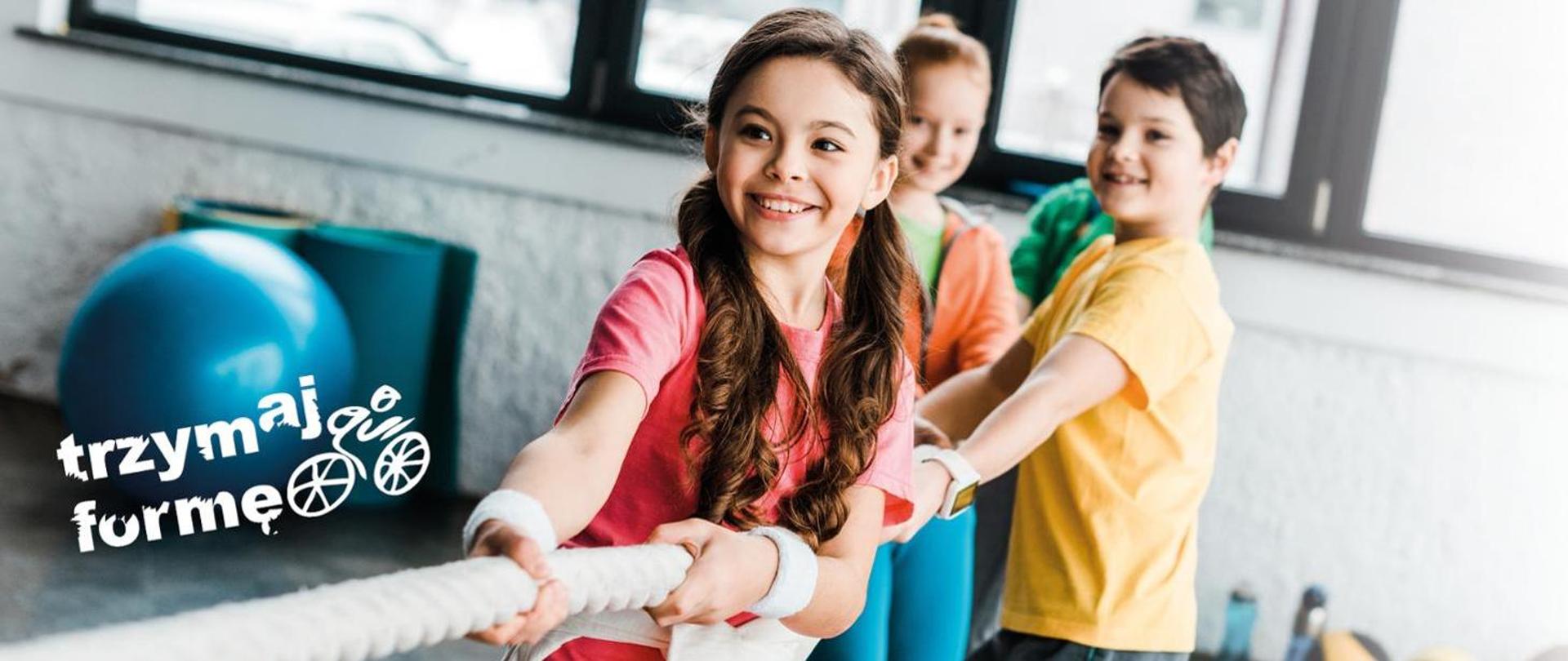 Na zdjęciu 3 dzieci przeciągających linę. Na pierwszym planie dziewczynka z długimi włosami w czerwonej koszulce, za nią chłopiec w żółtej koszluce, na końcu dziewczynka w pomarańczowej bluzie