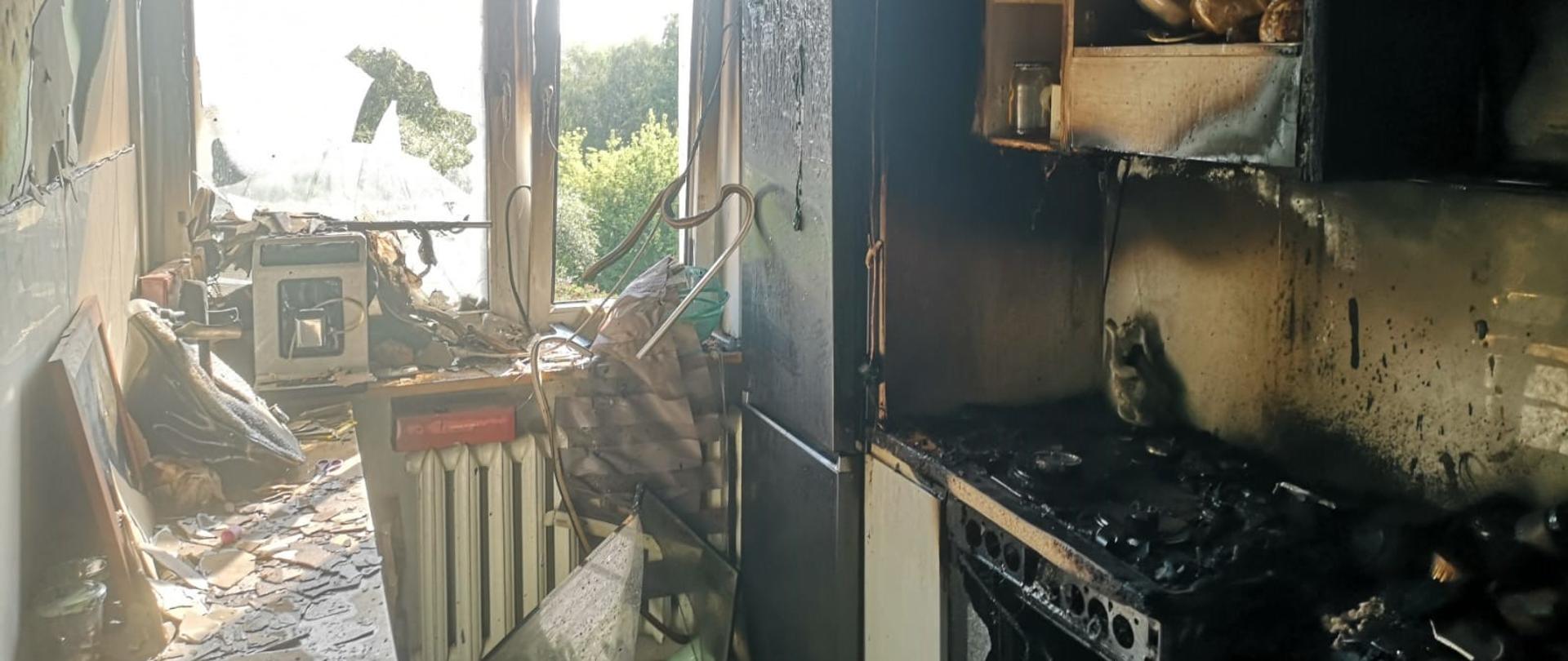 Zdjęcie przedstawia miejsce występowania pożaru, jest to pomieszczenie kuchenne wraz z całym jego wyposażeniem. Na zdjęciu po prawej stronie widzimy pozostałości po zabudowie kuchennej, zniszczoną kuchenkę gazową, tuż nad nią szafki na przybory kuchenne, które w skutek pożaru uległy okopceniu. Na środku zdjęcia widzimy okno, z powybijanymi szybami oraz stopionymi ramami. Po lewej stronie znajduje się stół na którym leży tynk ze ściany. Sufit pomieszczenia jest całkowicie czarny. 
