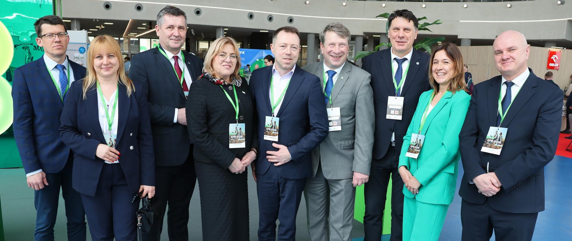 Grupa osób ubranych na elegancko w garnitury stoi w sali i pozują do zdjęcia. Na szyjach mają zielone smycze. 