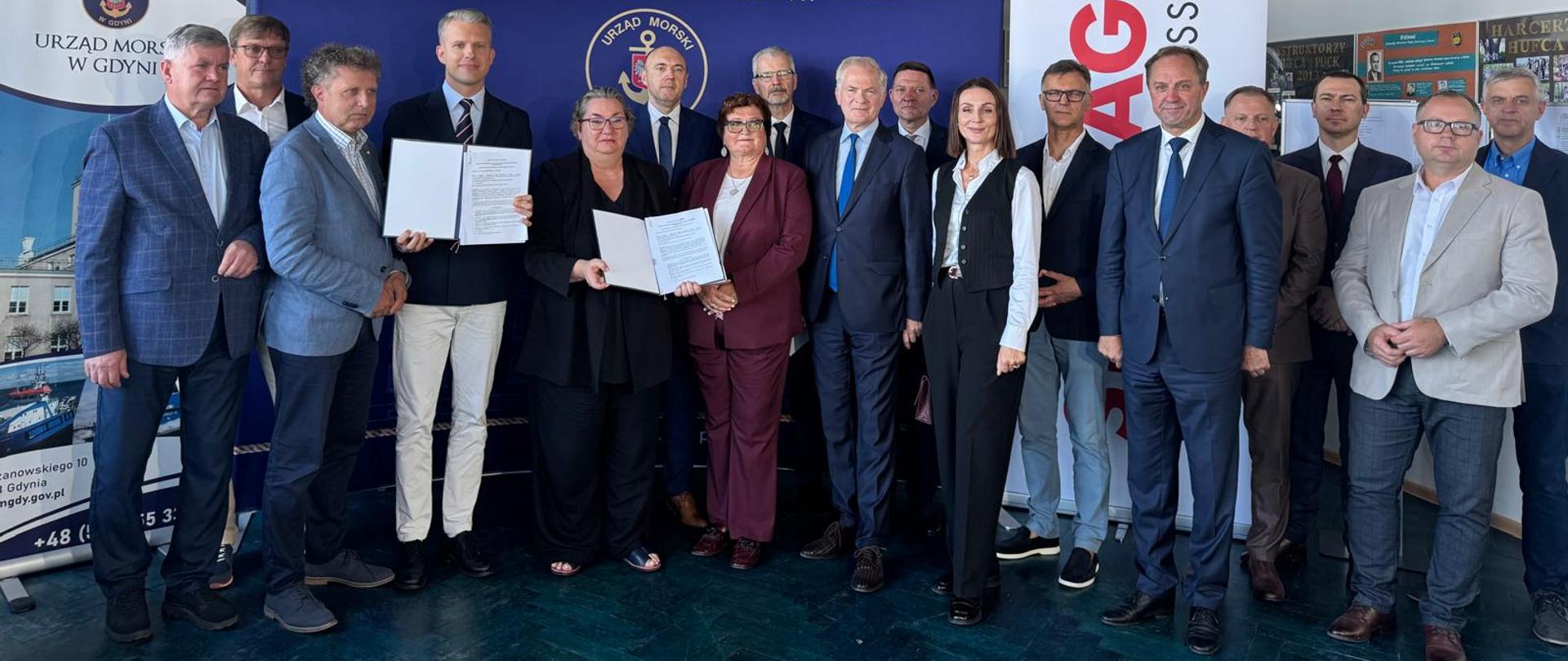 grupa osób stoi przed banerem z napisem urząd morski w Gdyni, dwie osoby trzymają w ręce podpisane wcześniej umowy