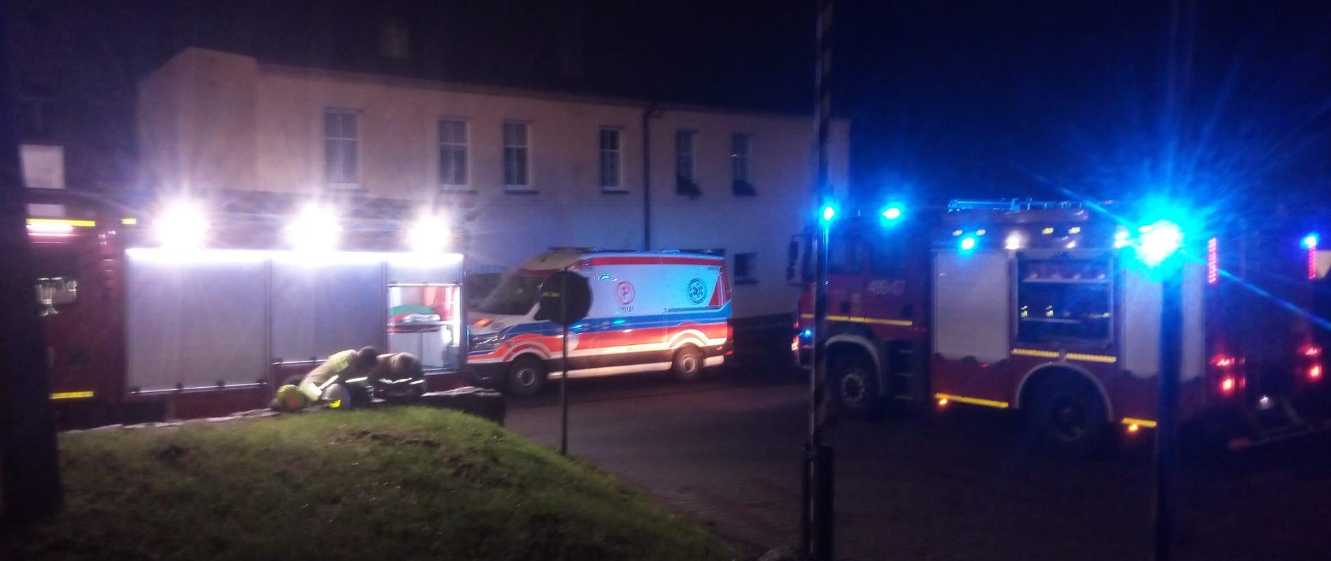 Na zdjęciu znajduje się karetka pogotowia, dwa wozy strażackie oraz budynek