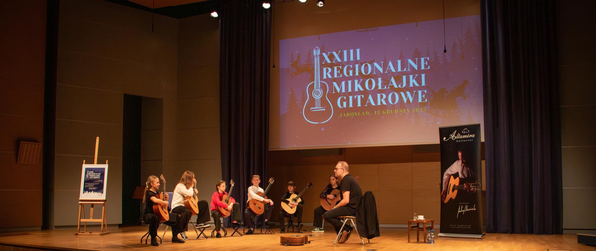 Szerokie ujęcie sceny, na której siedzi sześcioro dzieci z gitarami oraz dorosły mężczyzna. Na ekranie w tle duży napis "XXIII REGIONALNE MIKOŁAJKI GITAROWE JAROSŁAW, 12 GRUDNIA 2025". Na pierwszym planie widoczna publiczność siedząca w ciemnej sali.
