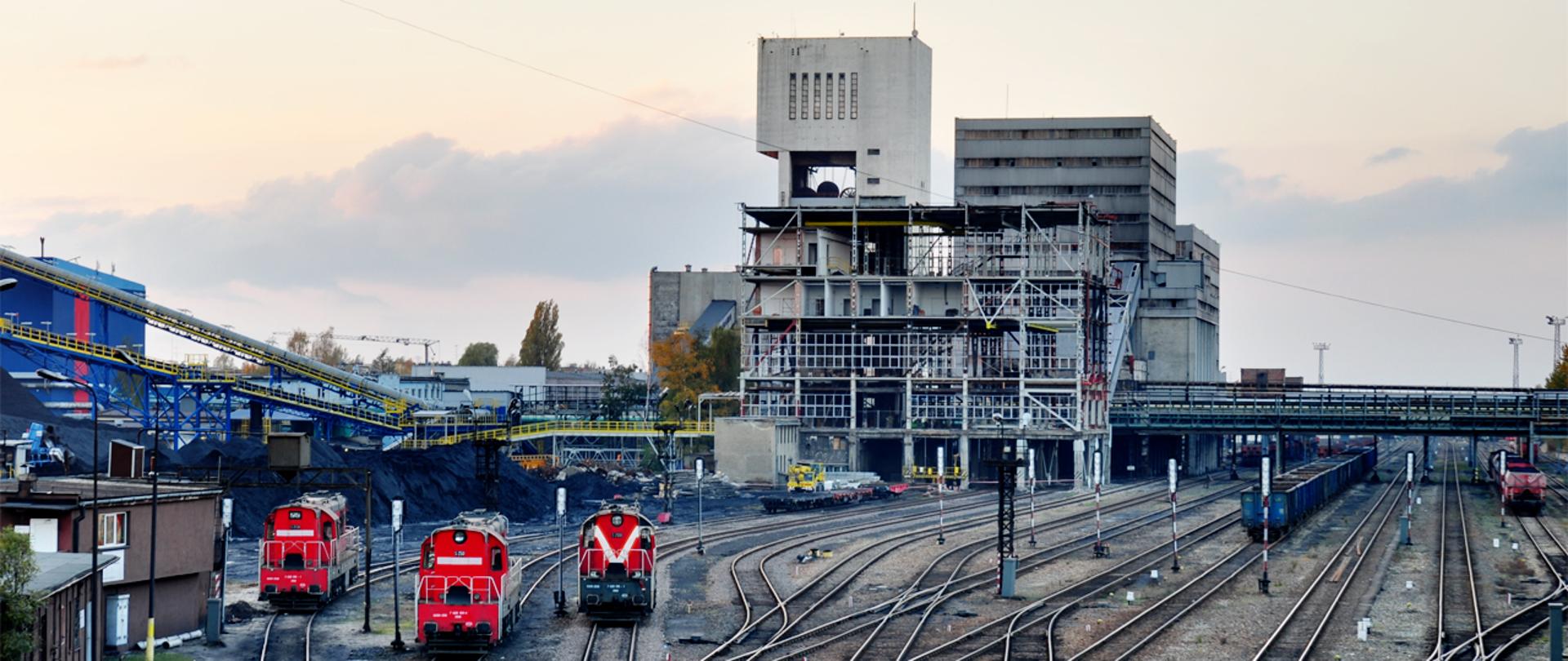Trzy pomalowane na czerwono lokomotywy stoją na torowisku przed wieżą szybową kopalni węgla kamiennego