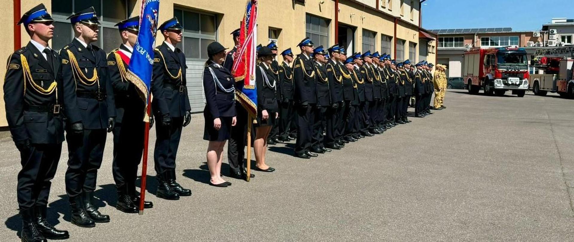 Fotografia wykonana podczas dnia strażaka w komendzie powiatowej Państwowej Straży Pożarnej w Mielcu. Na zdjęciu stoją funkcjonariusze komendy powiatowej PSP w Mielcu na placu przed jednostką ratowniczo-gaśniczą nr 2. Wszyscy strażacy stoją w postawie zasadniczej i są umundurowani w mundury służbowe i galowe. Na pierwszym planie stoi poczet sztandarowy wraz z sztandarem Komendy Powiatowej PSP w Mielcu.