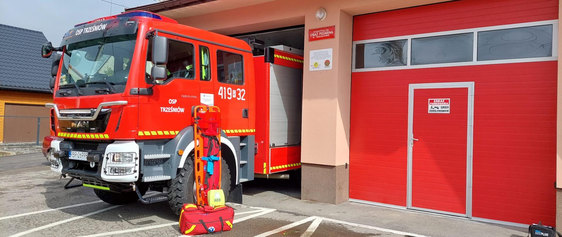 Kolorowa fotografia wykonana w pogodny dzień na zewnątrz strażnicy OSP Trześniów. Na pierwszym planie średni samochód pożarniczy częściowo poza strażnicą. Na drugim planie o samochód oparty jest sprzęt ratownictwa medycznego tj: szyny Kramera, defibrylator AED i torba R1.