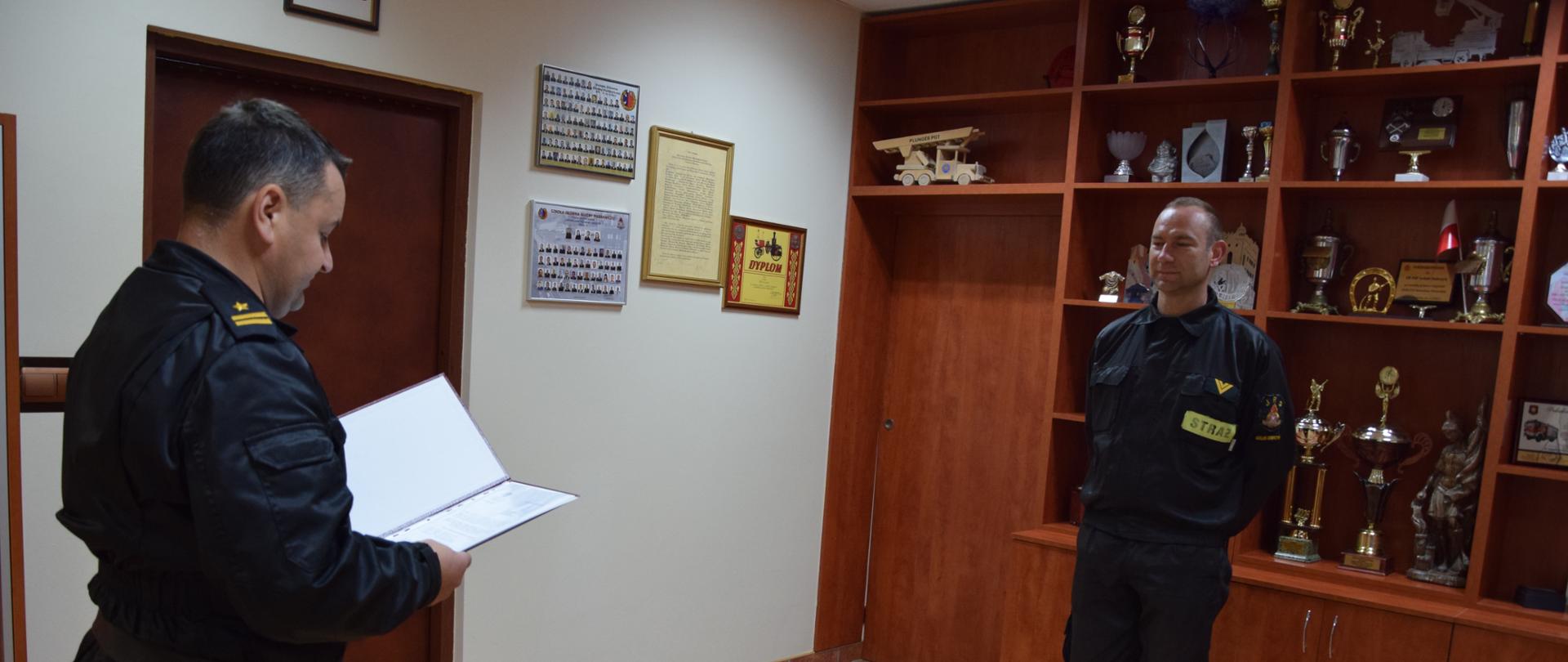 Dwóch strażaków stoi naprzeciwko siebie jeden z nich trzyma w dłoniach otwarta teczkę i czyta.