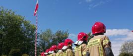Zbiórka strażaków w ubraniu specjalnym i hełmach na placu zewnętrznym komendy, strażacy widziani od tyłu, w tle na maszcie powiewa flaga Narodowa.