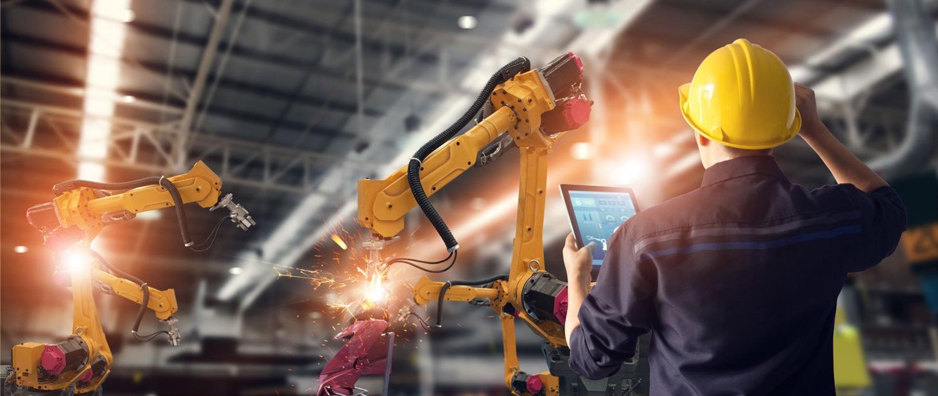 mężczyzna w kombinezonie i kasku z tabletem w dłoni w hali produkcyjnej ze spawającymi ramionami robotycznymi