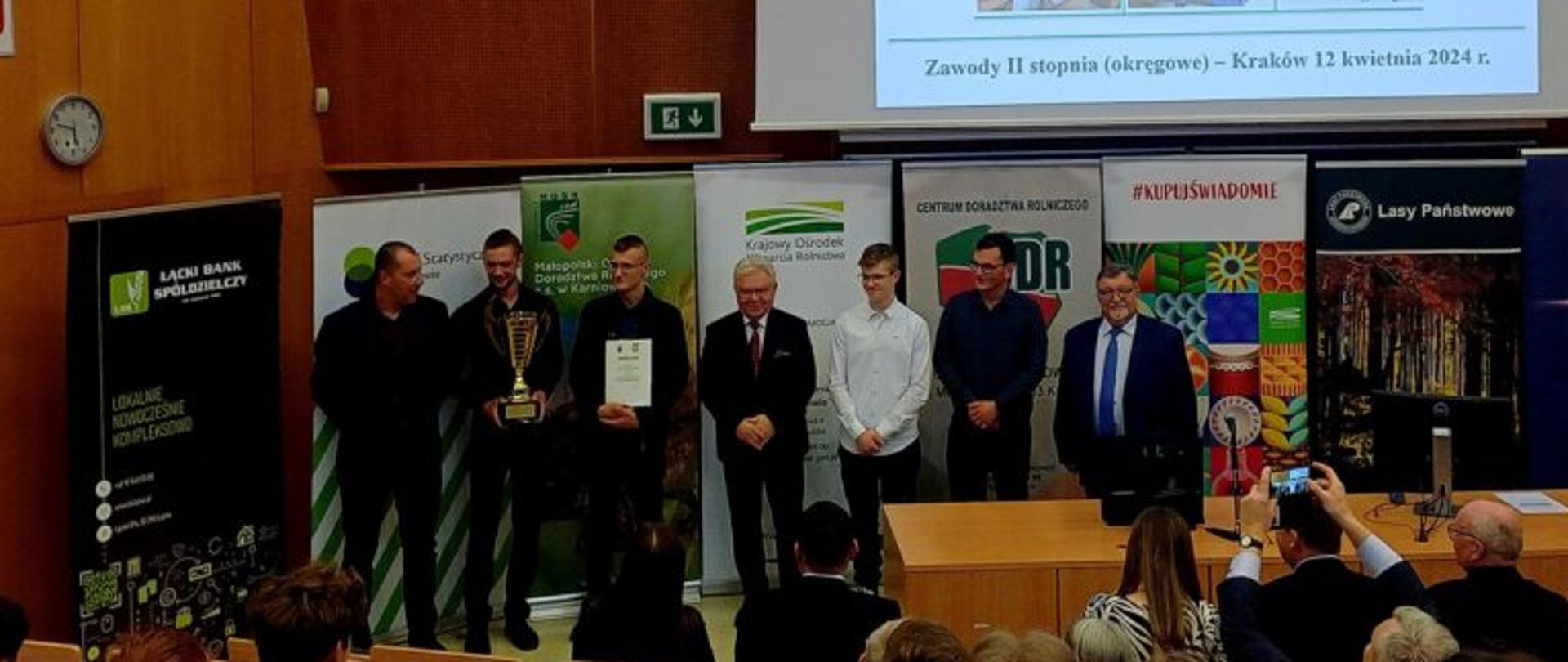 Uczestnicy i organizatorzy XLVII Olimpiady Wiedzy i Umiejętności Rolniczych na scenie podczas wręczenia nagród. W tle duży ekran z napisem „XLVII Olimpiada Wiedzy i Umiejętności Rolniczych, Kraków 12 kwietnia 2024 r.” oraz logotypy partnerów, m.in. Lasów Państwowych i Krajowego Ośrodka Wsparcia Rolnictwa. Jeden z laureatów trzyma złoty puchar.