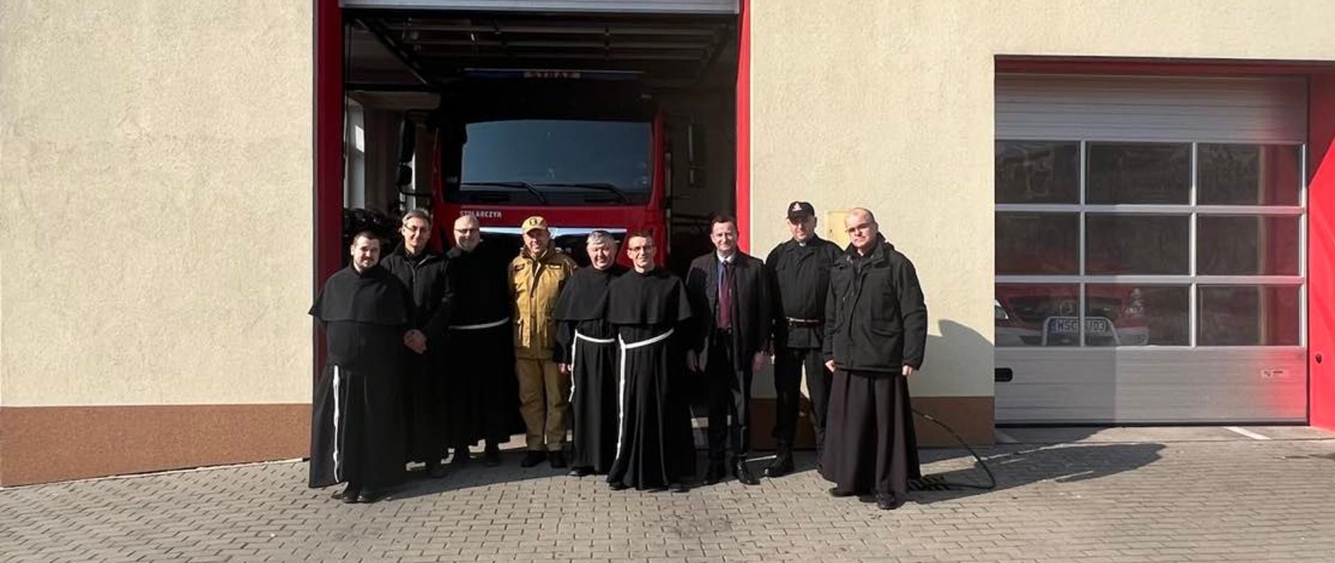 Pamiątkowe zdjęcie grupowe - mazowiecki komendant wojewódzki PSP nadbrygadier Jarosław Nowosielski w piaskowym mundurze służbowym stoi wśród zakonników w czarnych habitach, przepasanych białymi sznurami. Grupa stoi przed strażnicą OSP Niepokalanów - w uchylonej bramie widoczny samochód strażacki. Nad dwoma bramami widoczny herb OSP Niepokalanów i napis "OSP w Niepokalanowie"