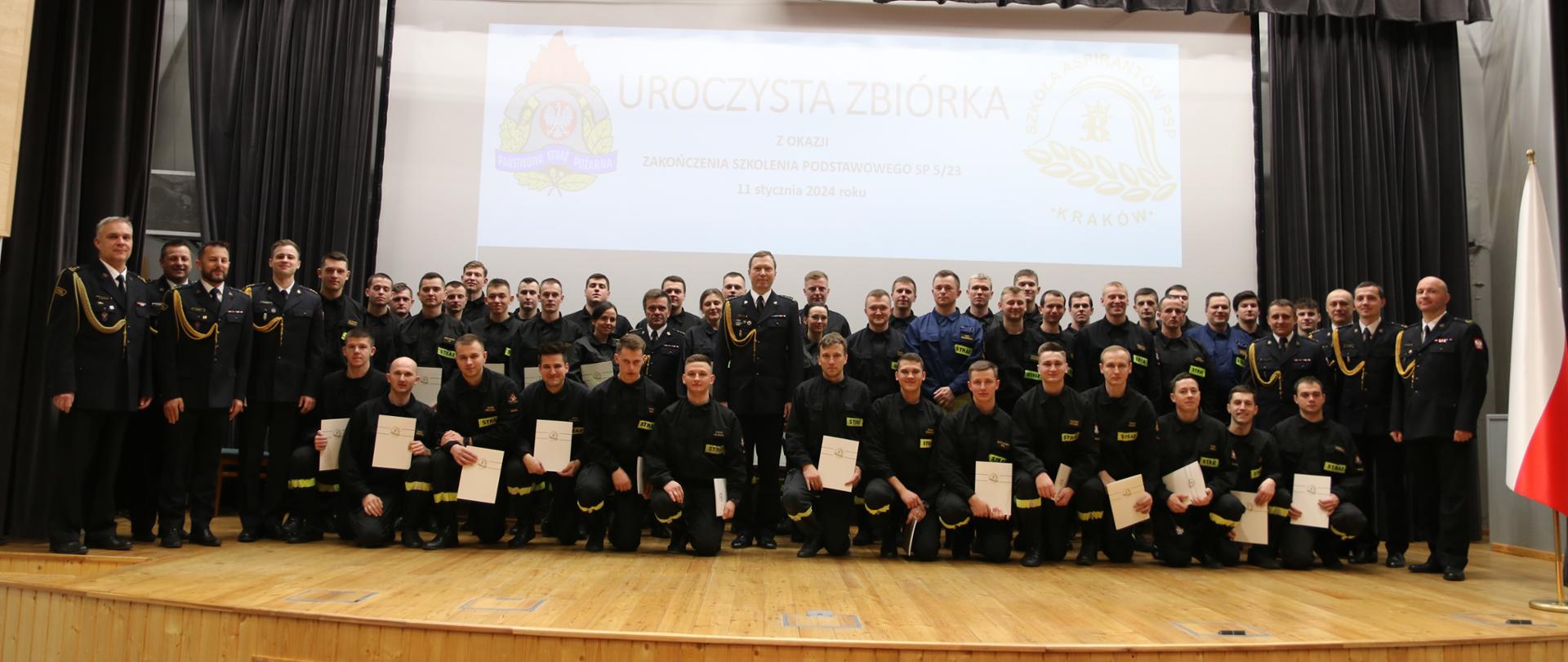 Pamiątkowe zdjęcie absolwentów szkolenia. Na scenie stoją trzymający w rękach teczki z dokumentami uczestnicy. Towarzyszą im z lewej: st.bryg. Robert Cieśla, z prawej st.bryg. Piotr Gancarczyk.