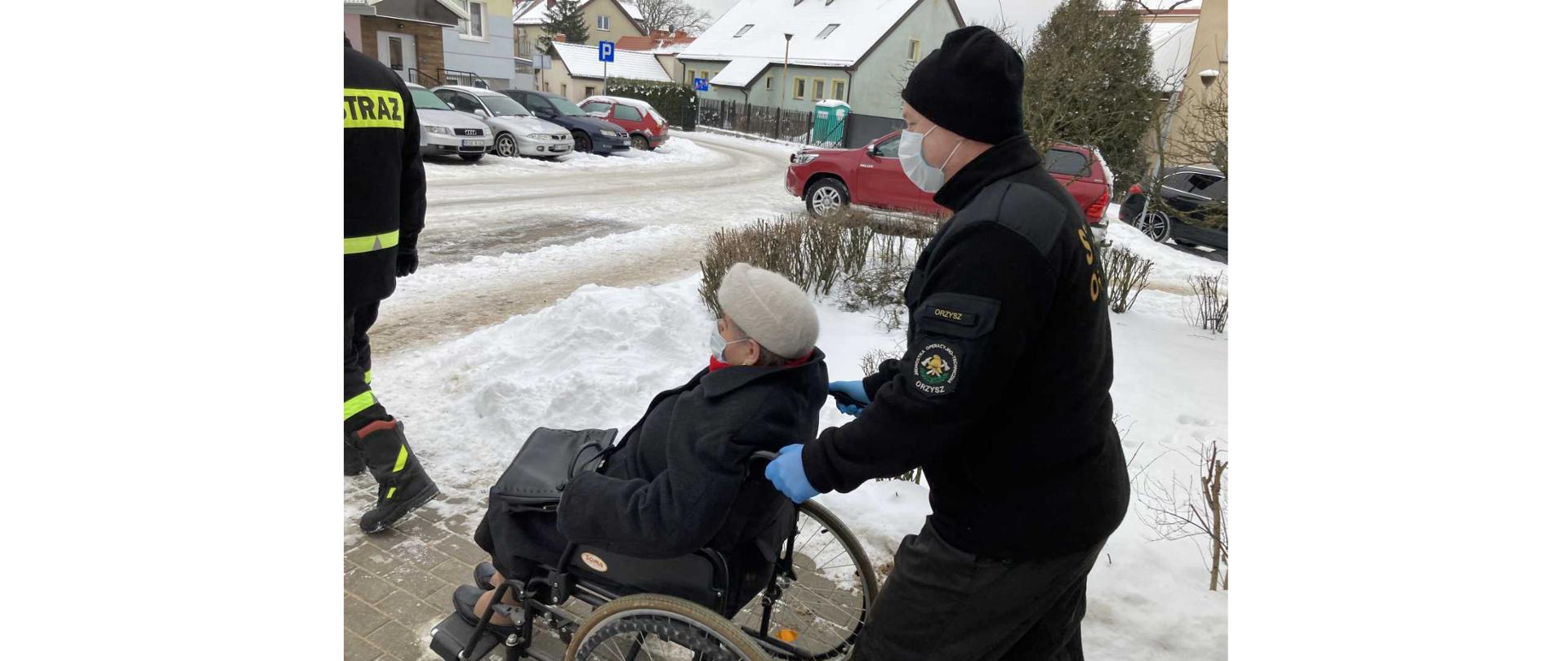 Kobieta na wózku inwalidzkim, pchanym przez strażaka.