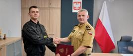Komendant Powiatowy Państwowej Straży Pożarnej w Człuchowie st. bryg. Radosław Batruch stoi w gabinecie i wręcza akt mianowania na stanowisko dowódcy zmiany st. asp. Karolowi Płóciennik.