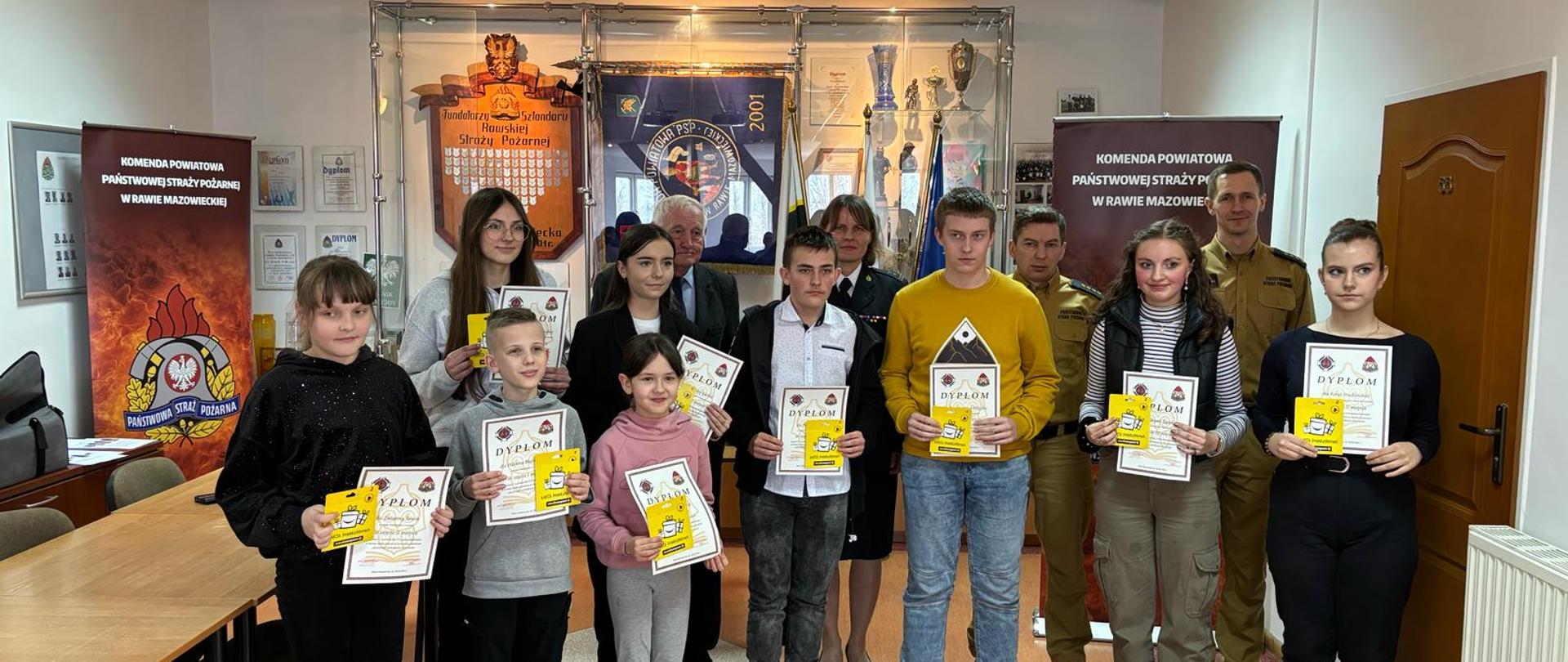 Na zdjęciu w dwóch rzędach stoją dzieci i młodzieży, laureaci konkursu oraz dorosłe osoby z czego trójka z nich jest w mundurach. Dzieci i młodzież trzymają w dłoniach dyplomy oraz nagrody.