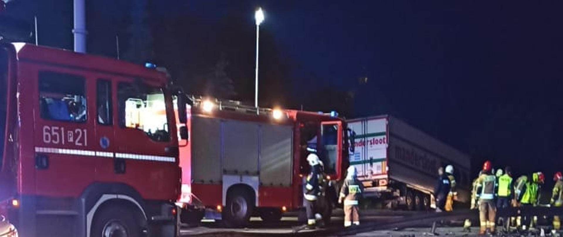 Zdjęcie przedstawia dwa samochody strażackie stojące na skrzyżowaniu. Obok stoi grupka strażaków. Za nimi widać przyczepę samochodu ciężarowego lezącego w rowie