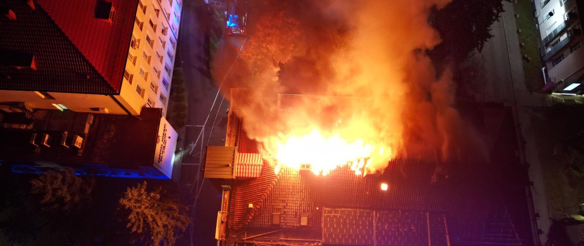Na zdjęciu widać dramatyczną scenę nocnego pożaru budynku. Ogień jest intensywny, jasnopomarańczowe płomienie trawią konstrukcję, a gęsty, ciemny dym unosi się wysoko w powietrze. Wokół budynku widać światła pojazdów pożarniczych, co świadczy o trwającej akcji gaśniczej. Ulice wokół są puste. Całość rozgrywa się w otoczeniu oświetlonych budynków szpitala.