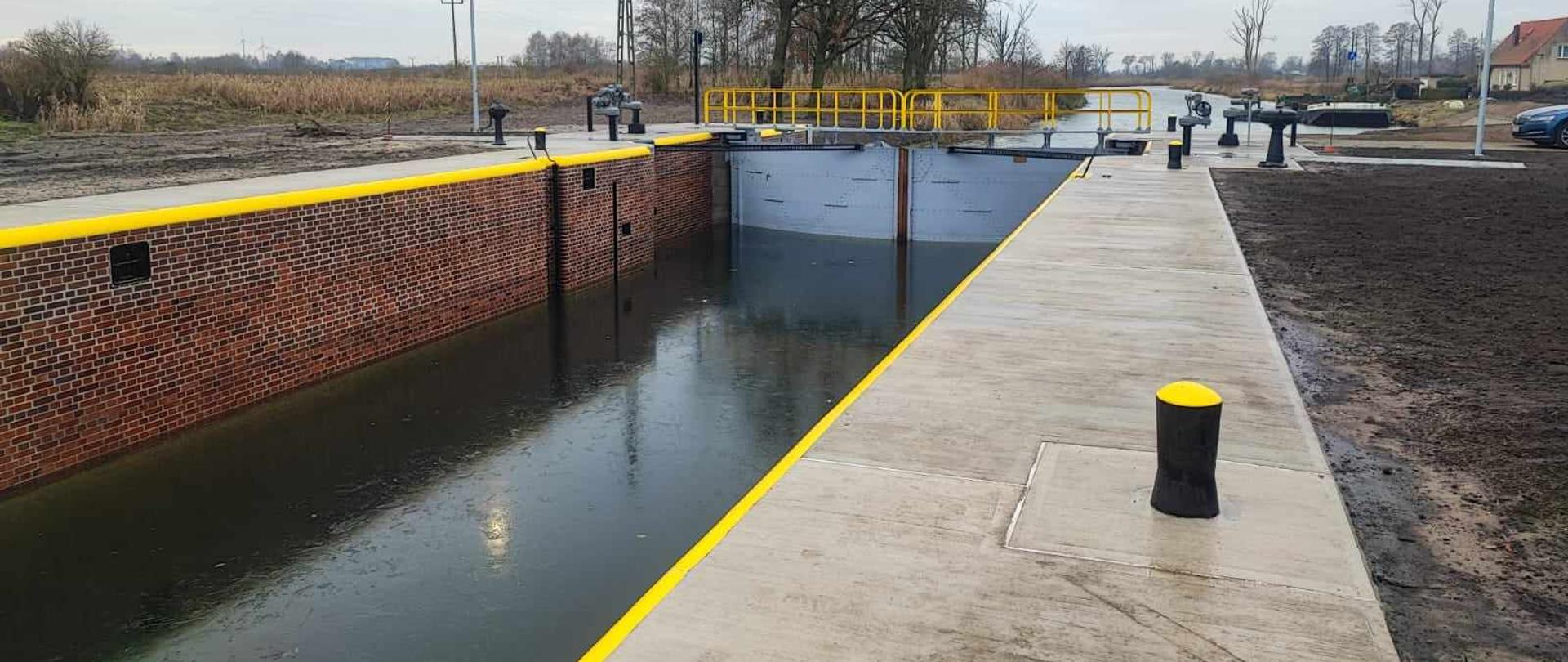  Śluza nr 8 Nakło Wschód po remoncie. Komora śluzy wypełniona wodą. Ściany z czerwonej, czystej cegły. Krawędzie świeżo malowane żółta farbą. Brzegi betonowe. Szare metalowe wrota śluzy. Wokół ponury jesienny dzień. Drzewa bez liści, suche łąki, goła ziemia. 