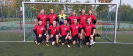 Zdjęcie grupowe drużyny z Białogardu. Strażacy stoją w bramce. Mają na sobie ubrania sportowe koloru czerwono czarnego. Jest ładny jesienny dzień.