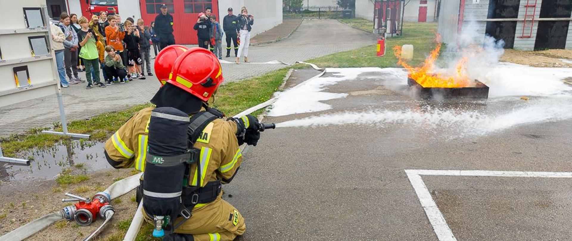 Na zdjęciu podczas pokazów strażackich dla dzieci i młodzieży widać dwóch strażaków gaszących pożar wzniecony w wannie z substancją ropopochodną. W tle grupa dzieci i młodzieży przyglądająca się gaszeniu pożaru. Zdjęcie wykonane za pleców strażaków na placu wewnętrznym komendy powiatowej PSP w Ostrowcu Św.