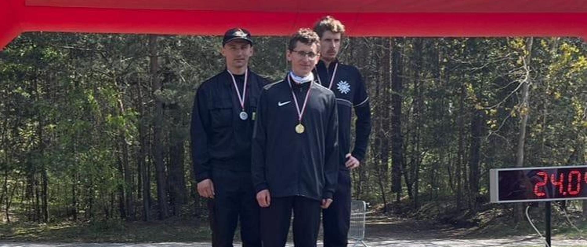 Zdjęcie przedstawia zwycięzców biegu przełajowego w kategorii do 35 roku życia stojących na podium po wręczeniu medali.
