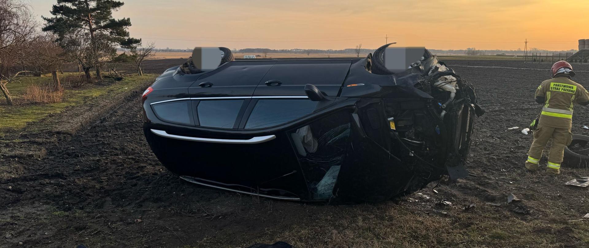 Na zdjęciu widać czarny samochód osobowy, który leży na prawym boku na polu. Przód pojazdu jest całkowicie rozbity. Obok samochodu stoi strażak w czerwonym hełmie i żółtym stroju z napisem Państwowa Straż Pożarna. Na polu porozrzucane są elementy pojazdu. W tle widać drzewa oraz pola. Jest widno, ale słońce zachodzi.