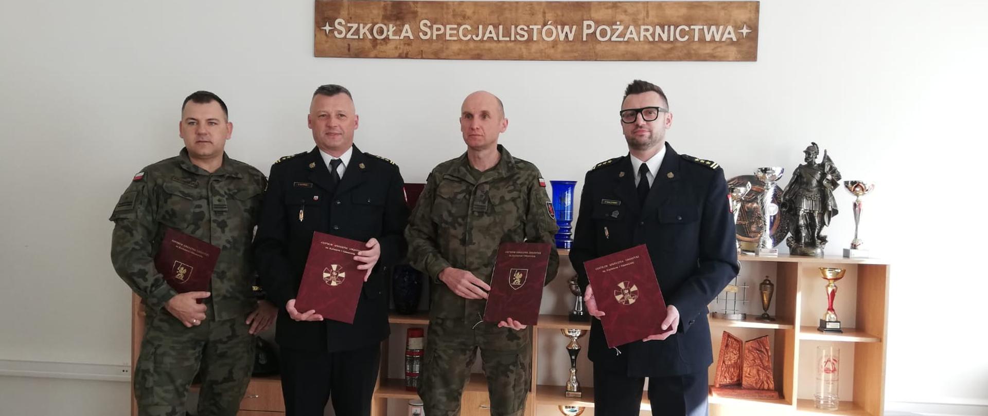 Stoją obok siebie i trzymają w rękach teczki z podpisanym uzgodnieniem od prawej starszy brygadier Piotr Tkaczyński Komendant Powiatowy Państwowej Straży Pożarnej w Świeciu, pułkownik Andrzej Dyk Komendant Centrum Szkolenia Logistyki w Grupie, starszy brygadier Robert Gutowski Komendant Miejski Państwowej Straży Pożarnej w Grudziądzu, major Dawid Kalinowski Komendant Szkoły Specjalistów Pożarnictwa w Grupie. W tle na ścianie wisi napis:"SZKOŁA SPECJALISTÓW POŻARNICTWA" oraz stoi gablota z pucharami i statuetkami.
