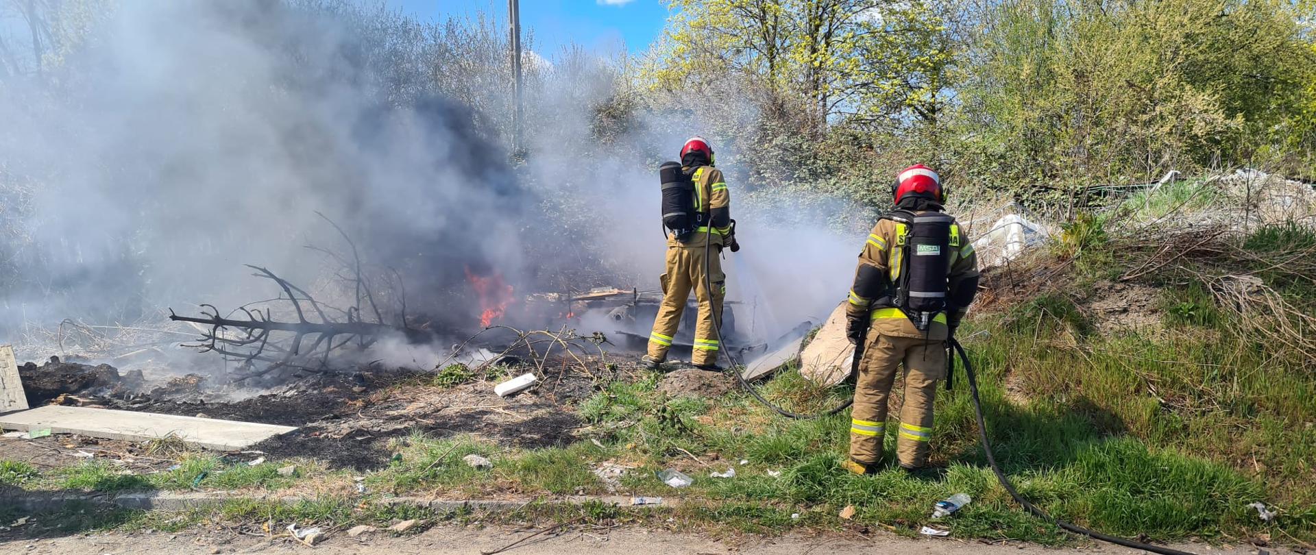 Podsumowanie zdarzeń w dniach 11.04 - 17.04.2022 r. - zdjęcie przedstawia 2 strażaków podających prąd wody na pożar dzikiego wysypiska śmieci