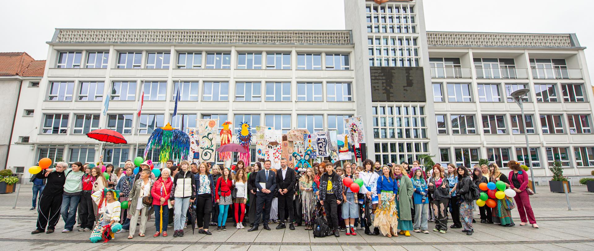 Zapytanie zostało przetworzone
Duża grupa osób, w tym uczniowie i dorośli, pozuje do zbiorowego zdjęcia na placu przed dużym, nowoczesnym budynkiem. Niektórzy trzymają kolorowe balony i artystyczne transparenty. W centrum grupy stoi dwóch mężczyzn w garniturach. Budynek ma wiele okien, a po prawej stronie znajduje się wysoka wieża zegarowa.