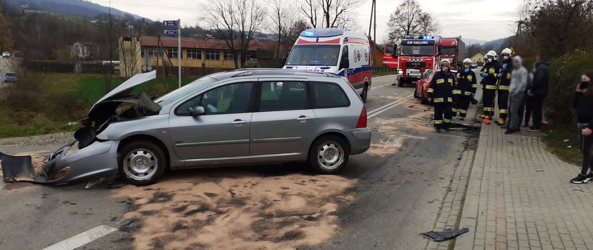Rozbity szary samochód. Plama oleju zasypana sorbentem oraz strażacy.