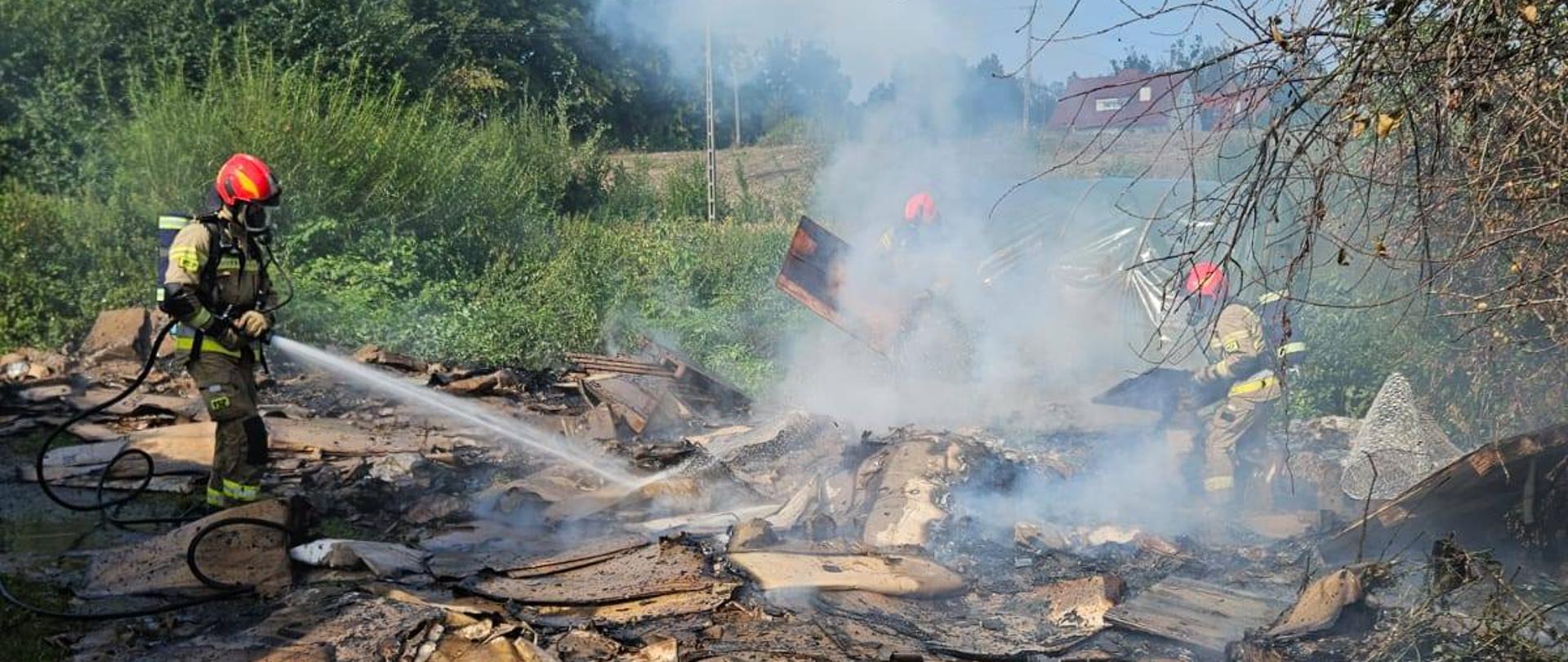 Strażacy w pełnym umundurowaniu pracują przy pożarze śmieci, niedopalone kartony . Unosi się dym. Pogodnie, słonecznie.