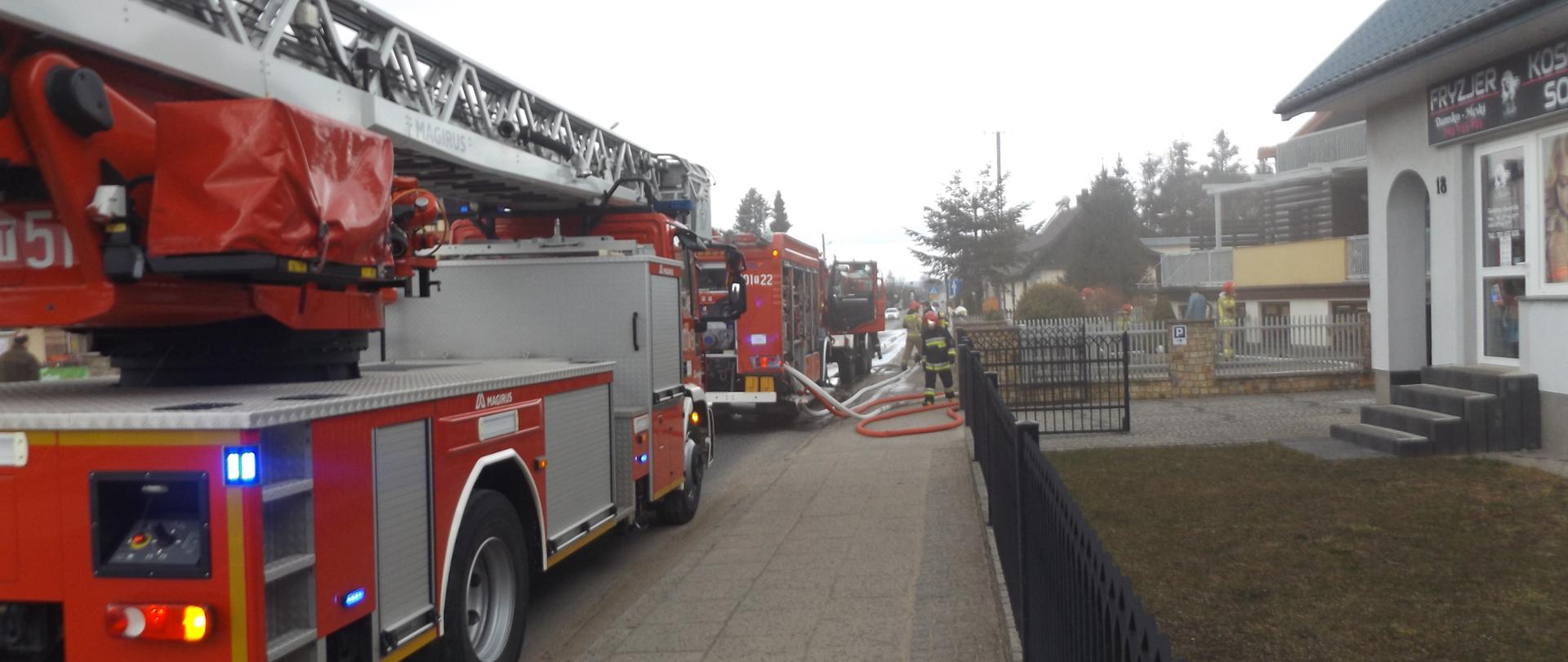 Zdjęcie przedstawia samochód pożarniczy - drabinę mechaniczną oraz inne samochody straży pożarnej stojące w oddali. Z prawej strony znajduje się wjazd na posesję na której doszło do pożaru. 