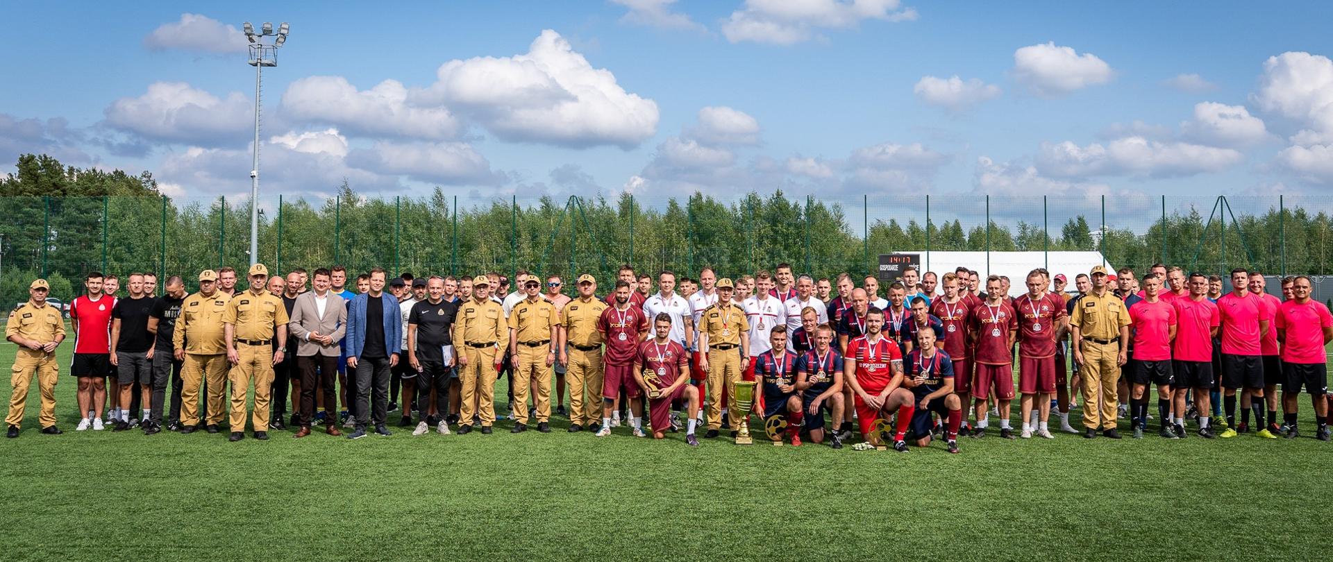 Duża grupa dorosłych mężczyzn stoi na trawiastym boisku w słoneczny dzień. Niektórzy z nich mają na sobie jednolite mundury w kolorze beżowym lub sportowe stroje w różnych kolorach.