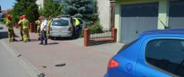 Masłowo, ulica Matejki. Samochód osobowy uderzył w ogrodzenie posesji. Na pierwszym planie fragment auta osobowego koloru błękitnego. Zanim samochód osobowy koloru srebrnego uczestniczący w kolizji , widziany od tyłu. Wokół niego strażacy i policjant oraz "cywil". W tle zabudowania i drzewa.