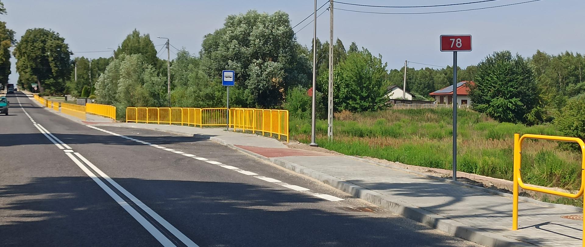 DK78 Kije - Chmielnik - jednojezdniowa droga asfaltowa, wzdłuż drogi chodnik, żółte barierki, w oddali zielone zarośla 