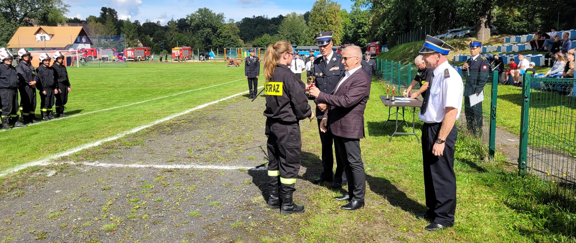 boisko sportowe, na bieżni boiska grupa strażaków podczas ceremonii wręczania pucharów po zawodach, druhna odbierająca puchar z rąk starosty, obok komendanci 